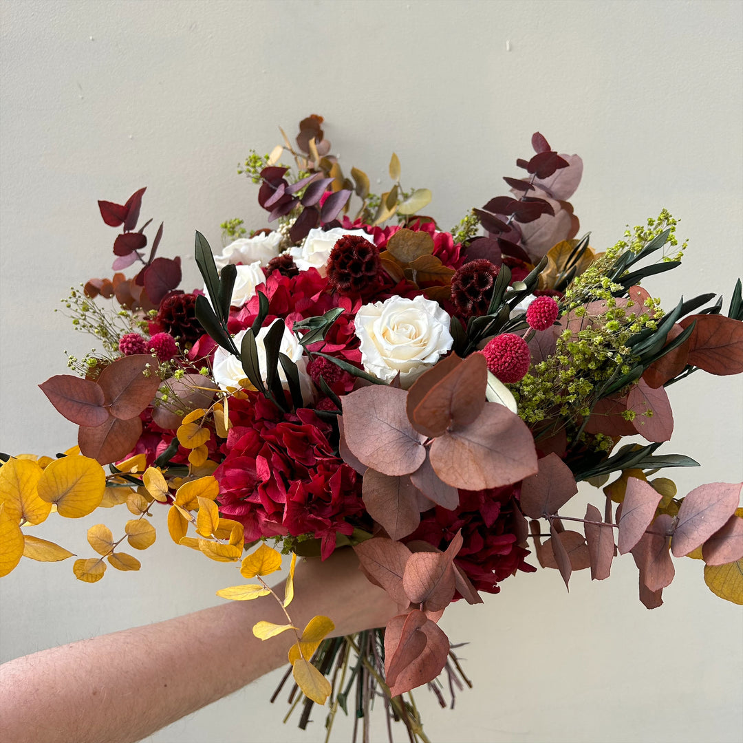 Un bouquet éternel tenu à la main, composé de roses blanches épanouies entourées de fleurs d'un rouge profond et de bordeaux, des baies, des hortensias préservés et des feuilles pourpres. Des touches de verdure et des herbes vert pâle ajoutent contraste et texture à l'ensemble dense et richement coloré